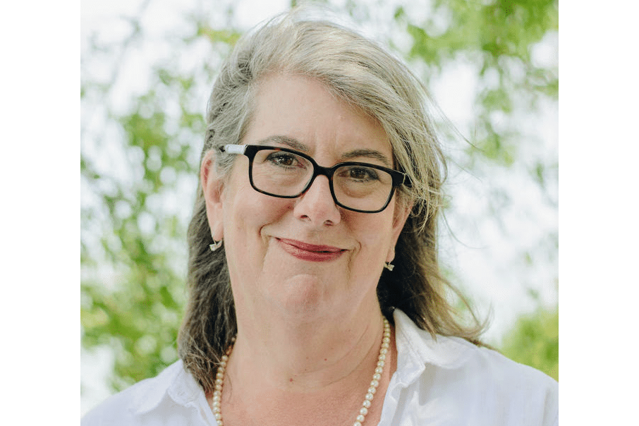 A woman wearing glasses and pearls smiles in front of trees.