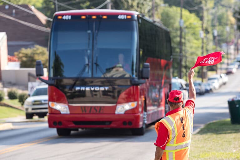person in orange vest and red hat directing a shuttle bus with red flags.
