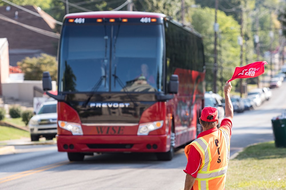 person in orange vest and red hat directing a shuttle bus with red flags.