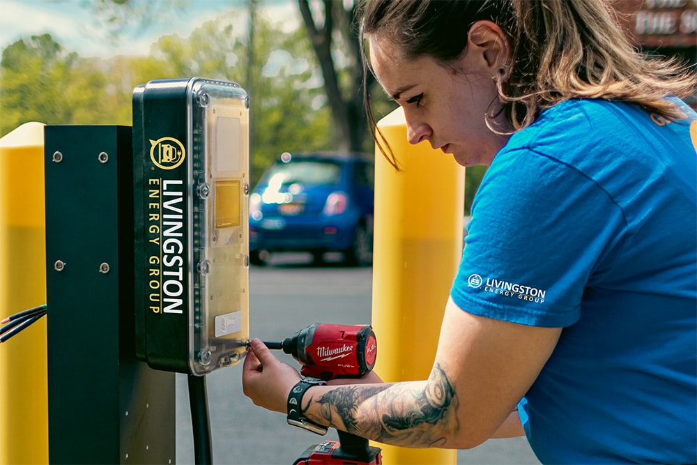 A woman wearing a blue T-shirt with a Livingston energy group logo on it, using a drill to install an EV charging station in a parking lot