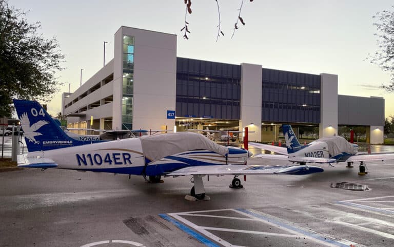 Two small aircraft with “emergency evacuation” markings parked in front of a modern multi-story parking garage at dusk.