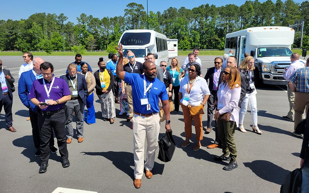 A group of people with badges listening to a man gesturing while standing in a parking lot with buses in the background.
