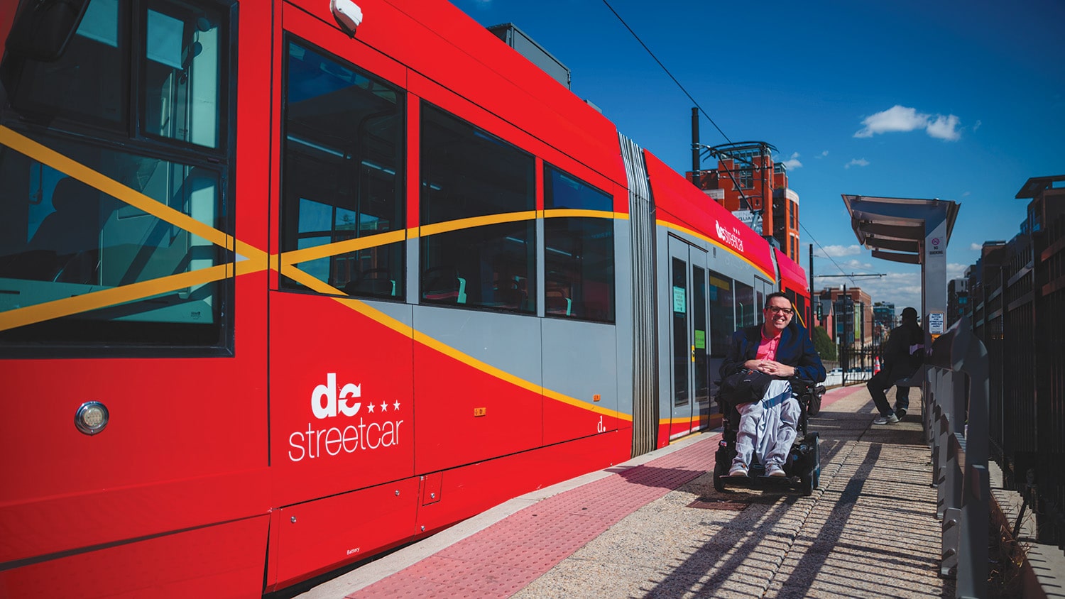A person in a wheelchair waits at a station beside a red streetcar on a sunny day.