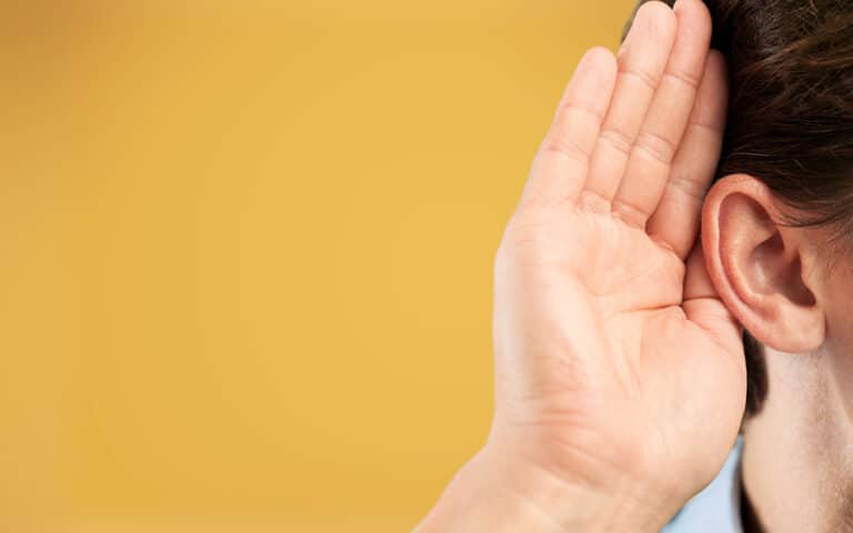 A person cupping their hand behind their ear to hear better, against a solid orange background.