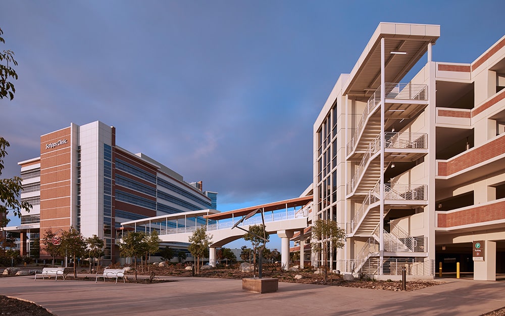 A modern hospital building with a pedestrian bridge connected to a multi-level parking structure at dusk.