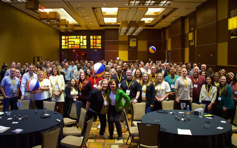 A group of people smiling at a conference hall with beach balls being tossed, some standing and some seated at round tables.