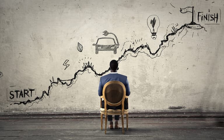 A man seated in front of a wall with a chalk drawing depicting a path from start to finish including symbols of a car, obstacles, and a lightbulb.