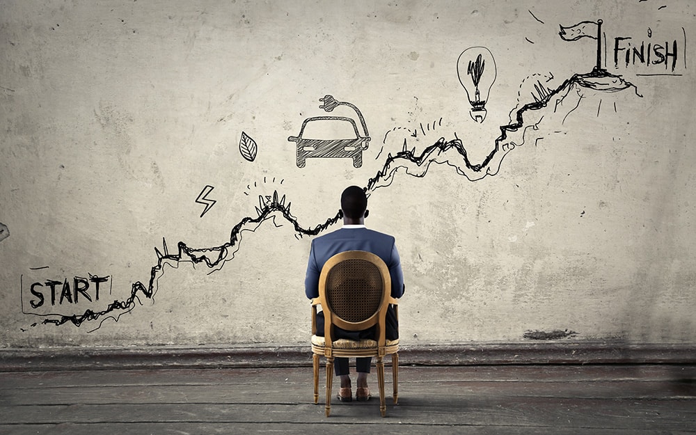 A man seated in front of a wall with a chalk drawing depicting a path from start to finish including symbols of a car, obstacles, and a lightbulb.