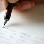A close-up of a hand holding a fountain pen, writing Auto Draft in cursive script on a white sheet of paper.