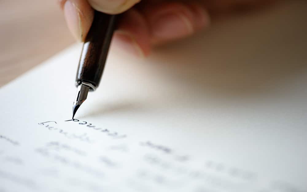 A close-up of a hand holding a fountain pen, writing Auto Draft in cursive script on a white sheet of paper.