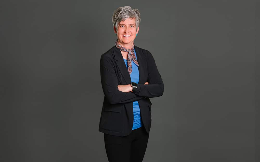 Professional middle-aged woman with short gray hair, wearing a black blazer and blue top, stands smiling with arms crossed against a CAPP-branded background.