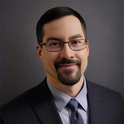 A professional headshot of a man with glasses wearing a business suit and tie.